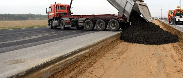 LKW schüttet Teer auf einen Weg, der zu einer Straße ausgebaut wird.