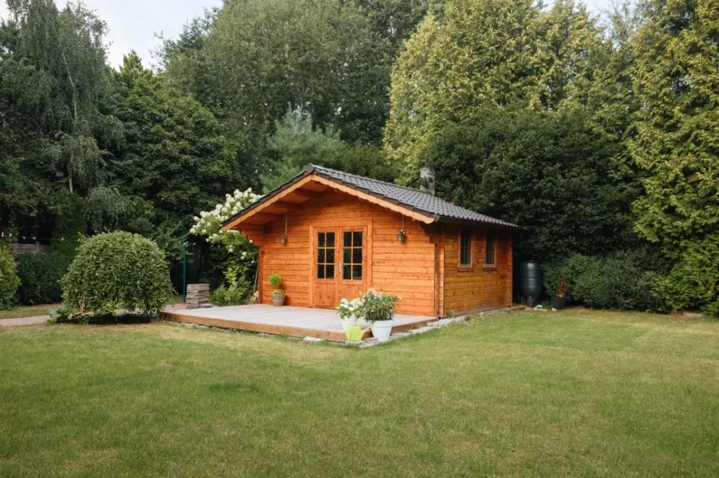 In einem Garten steht ein Gartenhaus aus Holz mit einem dunklen Dach und einer Terrasse davor