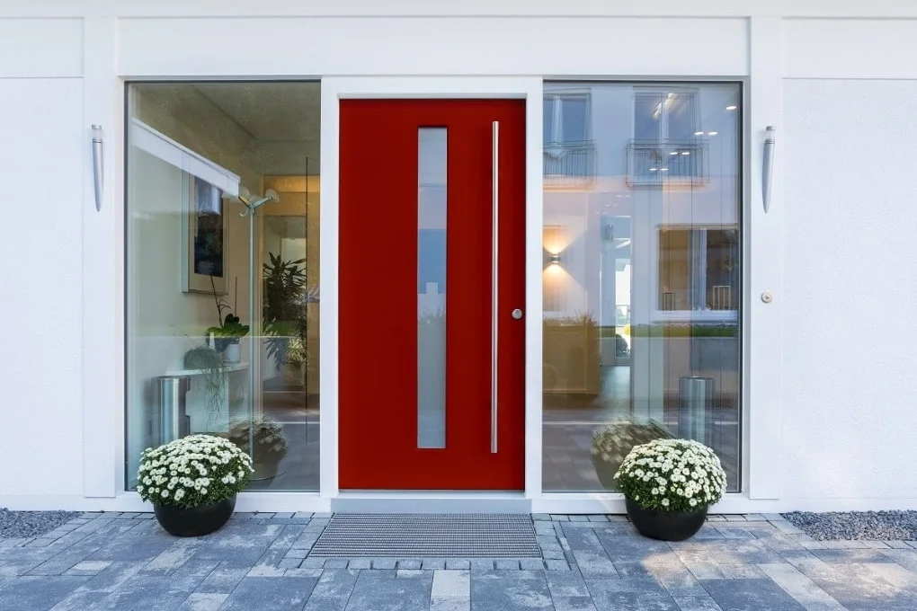 Eine Frontansicht vor einer roten Haustüre, die von zwei Glasfronten umschlossen ist.