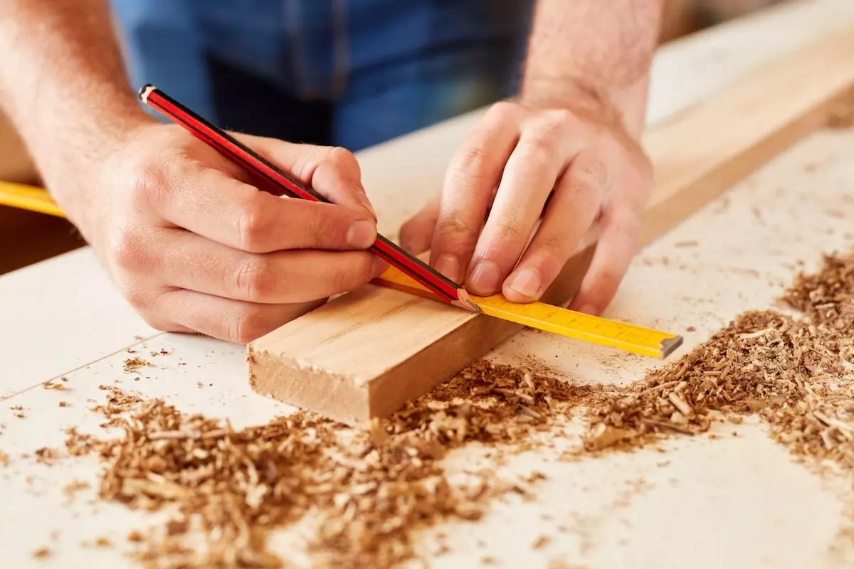 Eine Person arbeitet mit Holz und zeichnet mit Hilfe eines Bleistiftes und Meterstab eine Kennzeichnung auf ein Holzbrett.